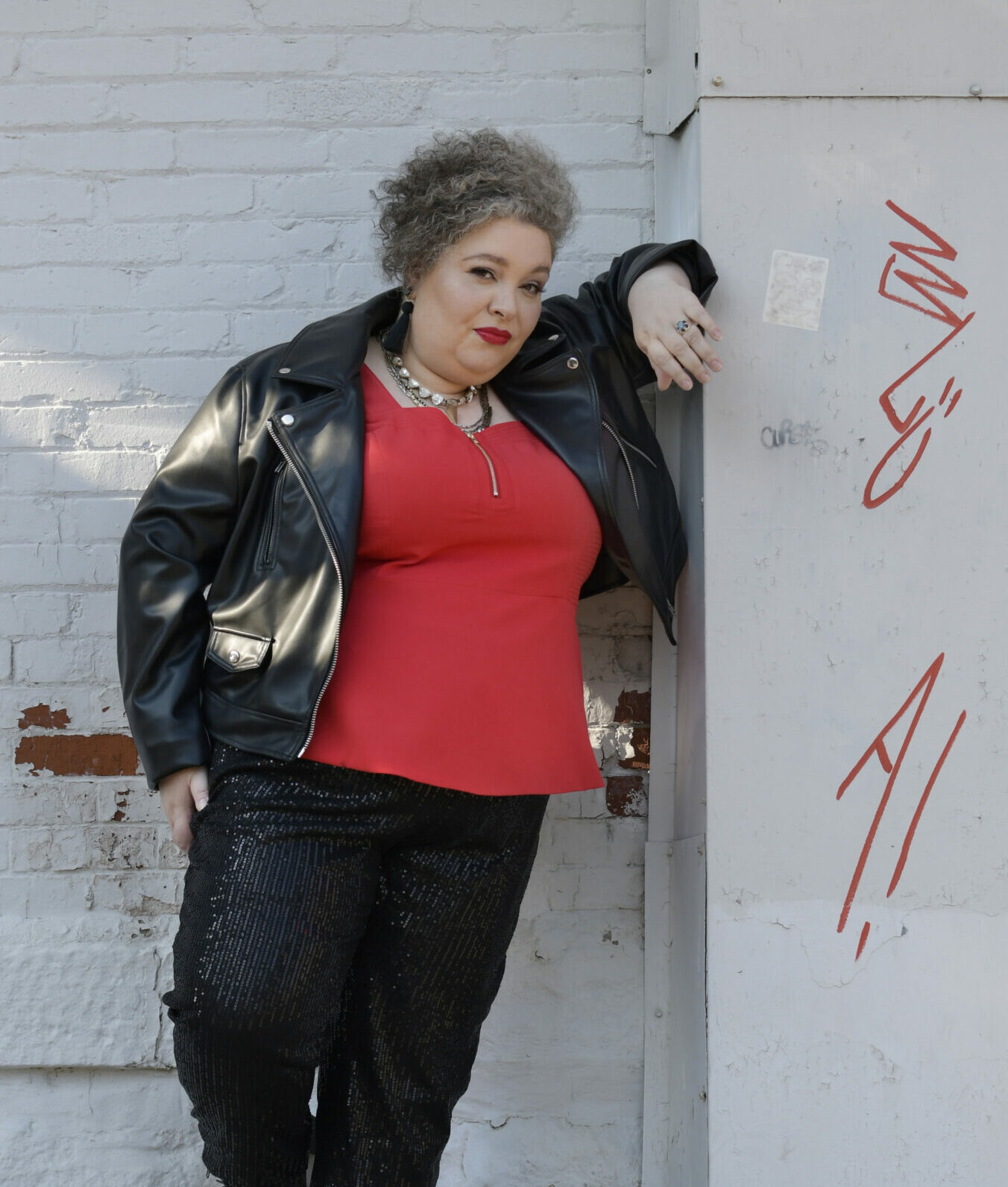 Lisa is dressed in a red tank top with black, sequined pants, a black leather jacket, and black boots. She is leaning against a brick wall with graffiti on it.