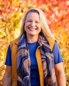 Anke Johnson is wearing a blue t-shirt and fall vest jacket. She has long blonde hair and is smiling into the camera.