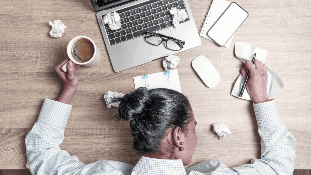 a woman is passed out, head on desk, hot cup of coffee in hand. There are scattered notes, eye glasses, an open laptop, and other items on the desktop. 