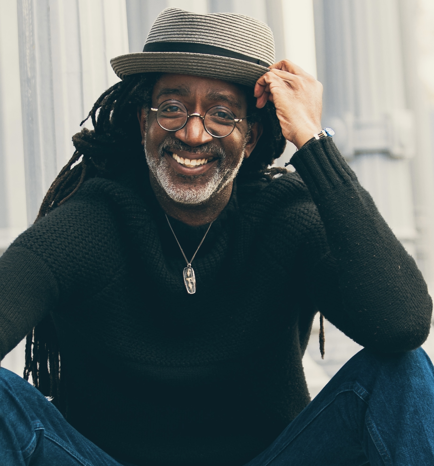 Conroy is wearing a goddess medallion, a dark shirt, and a grey fedora with a black hat band. He has long dark locs, glasses, and is smiling into the camera.