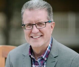 Jeff has short, salt-and-pepper hair, is wearing a plaid shirt, grey jacket, glasses, and is smiling at the camera.