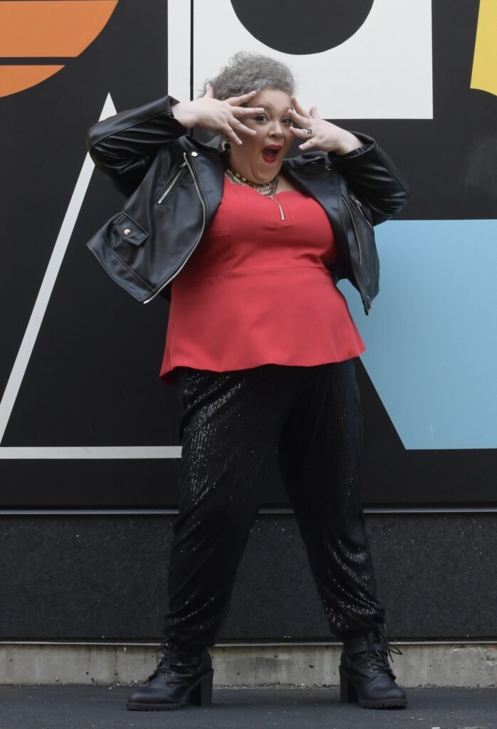Lisa's wearing a black leather jacket over a red tank top and black sequined pants. She's got her hands in front of her face, in a "jazz hands" style pose.